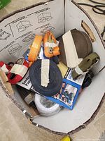 Box filled with rolled and loose tie-down straps in orange, blue, black, and green, with some packaging damage and partial visibility of product labels.