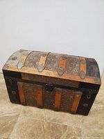 Closed domed trunk chest showing aged wooden slats, metal accents, and black leather or treated canvas panels.