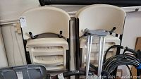 Set of four beige folding chairs stacked and folded, leaning against wall, with some surrounding garage items visible.