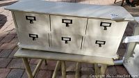 Front view of white wooden organizer with six drawers, placed on wooden bench table outdoors on brick pavement.