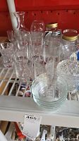 Mixed clear glassware including glasses, decanter, jars with metal lids, bowls, and covered candy dish placed on a white shelf with a red background, all needing cleaning.