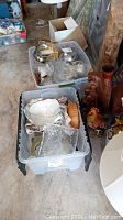 Two large plastic storage bins filled with various glass dishes, bowls, and barware, along with a carved vase and rooster figurine on the floor next to the bins