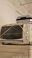 Oster toaster oven with two control dials on the front panel, metallic finish with a glass door and a cord wrapped around it.