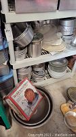 Full view of two storage shelves containing various aluminum pans, mixing bowls, baking tins, and the chicken cooker pot with lid.