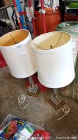 Two glass lamps with square bases, gilded trim and white linen shades. Both lamps have cords coiled at bases. One shade has slight imperfection (small dent). Lamps sit on concrete floor with various surrounding items.