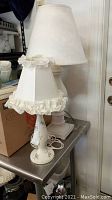 Photo showing two lamps on a metal table, including a small ceramic lamp with floral design and ruffled fabric shade, and larger lamp with wooden base and tall plain white shade.