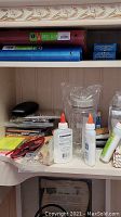 Shelves in cabinet containing binders, packs of staples, a pair of glasses, white glue bottles, and other small office items.