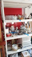 White plastic shelf unit with four shelves, containing various glassware, dishes, and miscellaneous items on the shelves. Red background and some other household items visible.