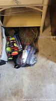 Photo showing tennis rackets in carrying bag and multiple cans of tennis balls placed on the floor under a table.