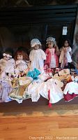 Five American Girl dolls seated in a row with an array of doll clothes and accessories laid out in front, showing the variety in clothing and doll appearance.