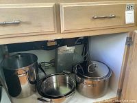 Large Calphalon stainless steel pots and pans including a large 5-gallon pot with lid, a frying pan, and a saucepan inside a cupboard.
