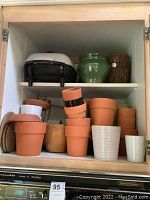 Photo of wooden kitchen cabinet shelf holding black and white electric countertop grill and various ceramic and terracotta pots.