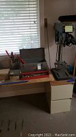 Full view of drill press standing on a two-drawer beige metal cabinet with table surface. Grey and red toolbox to the left containing wrenches, sockets, and other hand tools. Red bolt cutters also visible.