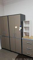 View showing two tall tan garage cabinets with metal handles and gray trim along with the shorter drawer cabinet topped with shelving unit.