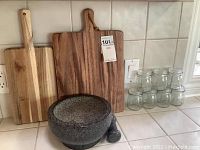 Photo of the granite mortar and pestle on a tiled kitchen countertop with three wooden cutting boards behind and six glass jars on the right side.