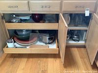 Wide view of lower kitchen cabinet with drawers and doors open showing various kitchenware items including plates, bowls, and appliances.