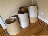 Overall view of six assorted lampshades in various neutral colors stacked on wooden floor against wall.