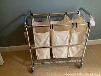 Metal linen cart with tubular silver metal frame, wheels, three cream cloth bags hanging from black hooks and wire shelf.
