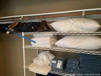 View of wire shelving unit containing white pillows, folded white and blue towels, fitted sheet, and hangers on rod above.