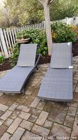 Two grey resin wicker chaise lounges on stone patio beneath tree and next to garden