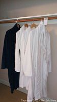 Photo showing five robes on hangers, four white and one dark blue, hanging on a wooden rack against a gray wall.