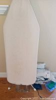 Ironing board with white fabric cover and metal legs folded underneath, standing upright.