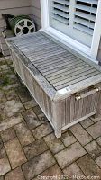 Wooden outdoor storage box with slatted top and brass-tone side handle, placed on a stone patio.