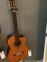 Full view of a natural wood Takamine acoustic guitar leaning against a wall, showing the body, neck, and headstock with tuning pegs.