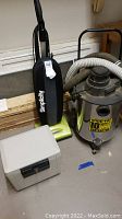 Photo showing all items in the lot: Simplicity upright vacuum, Shop-vac 10 gallon wet/dry vacuum, green handheld vacuum, and a gray tabletop safe.