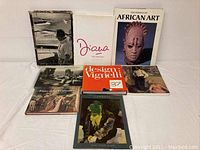 Seven art and photography books arranged on a white surface, titles include Diana The Portrait, Treasures of African Art, Design Vignelli 37, Pierre della Francesca, and two other art books.
