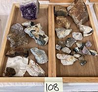 Photo showing a large purple amethyst crystal cluster and multiple assorted mineral rock specimens in a wooden tray.