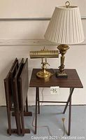 Four wooden nesting tables with a brass table lamp with white pleated shade and a brass desk lamp on top.
