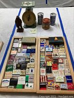 Wooden trays displaying a large assortment of collectible matchbooks and vintage tins, two Polar soda cans, the cast iron fisherman figurine, and the Universal metal scale on a table with a white covering.