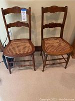 Two antique wooden chairs with cane seats. Both chairs have carved backrests and turned legs. One chair has an intact cane seat; the other shows cane damage.