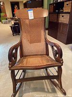 Front view of solid wood rocking chair with intact caned seat and backrest, scalloped back top rail and scrolled armrests.