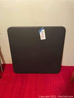 Black square card table leaning against a wall on a red carpeted floor. Top surface is visible with a tag attached.