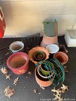 Collection of various sized clay and ceramic pots stacked together, with green wooden birdhouse on top and a green extension cord coiled on the side, plus metal hanging baskets behind.