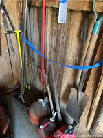 Photo showing garden tools arranged against wooden shed wall including shovels, rake, saw, and hedge clippers.