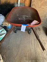 Front view of red metal wheelbarrow basin with a tag attached to the front edge and wooden handles visible