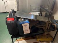 Wide view showing Bosch mitre saw mounted on black metal stand, with attached lot label and part of a Black & Decker sander case visible.