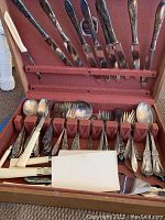 Opened case showing a variety of silver flatware consisting of knives with dark and white handles, forks, spoons, and serving utensils organized in a lined case.
