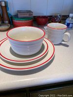 Photo showing stacked dinner plate, salad plate, bowl, and a large mug with white ceramic and red trim on a kitchen counter.