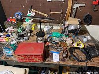 Wide view of work table showing a variety of hand tools, a red metal toolbox, boxes, and electrical components scattered across the surface.