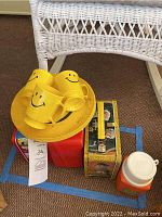 Photo showing yellow smiley face plastic cups and plate, red Peanuts lunchbox, Muppet Show lunchbox, and orange thermos grouped under white wicker furniture.