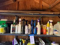 Wide shot of wooden shelves displaying assorted plastic spray bottles, jugs, and containers with yard and garden treatments in a shed environment.