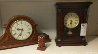 Two wooden quartz mantel clocks and one small wooden candle holder displayed on a shelf. One clock has a curved top with decorative inlays, the other has a rectangular frame with columns and visible pendulum.