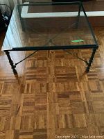 Square coffee table with clear glass top and black metal frame standing on parquet wood floor. Includes turned metal legs and metal cross supports in X shape underneath the table.