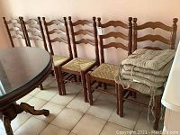 Full view of six wooden ladder back dining chairs with woven rush seats. Four chairs are visible entirely, two partially, aligned against a wall next to a round wooden table.