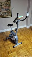Manual exercise bike with grey and silver frame, black seat, pedals with straps, dual handlebars, and display unit.