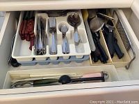 Photo showing drawer with flatware including forks, spoons, steak knives with wooden handles, and additional kitchen tools like a mesh strainer, scoops, peelers.
